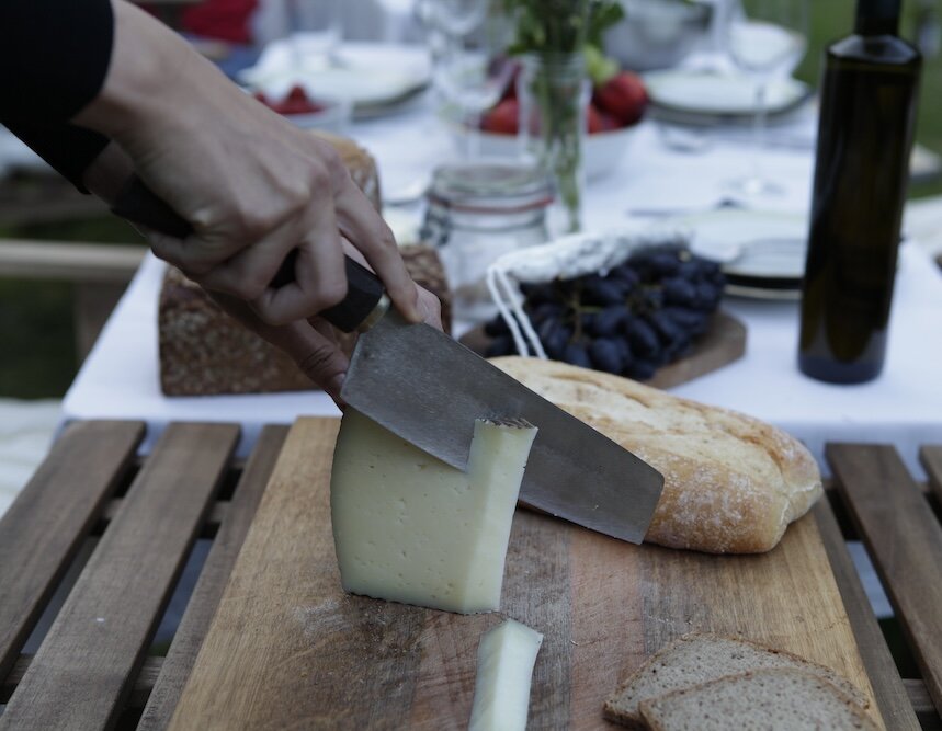 Käsestück wird mit einem großen Messer geschnitten, daneben liegen Brotlaibe